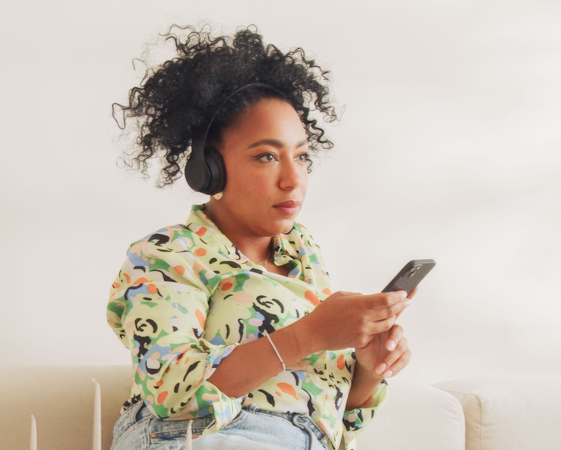 Eine Frau mit lockigem Haar trägt Kopfhörer und hält ein Smartphone mit der nuumi-App in den Händen, während sie auf einem beigen Sofa sitzt. Sie trägt ein farbenfrohes Hemd mit abstraktem Muster und schaut konzentriert in die Ferne.