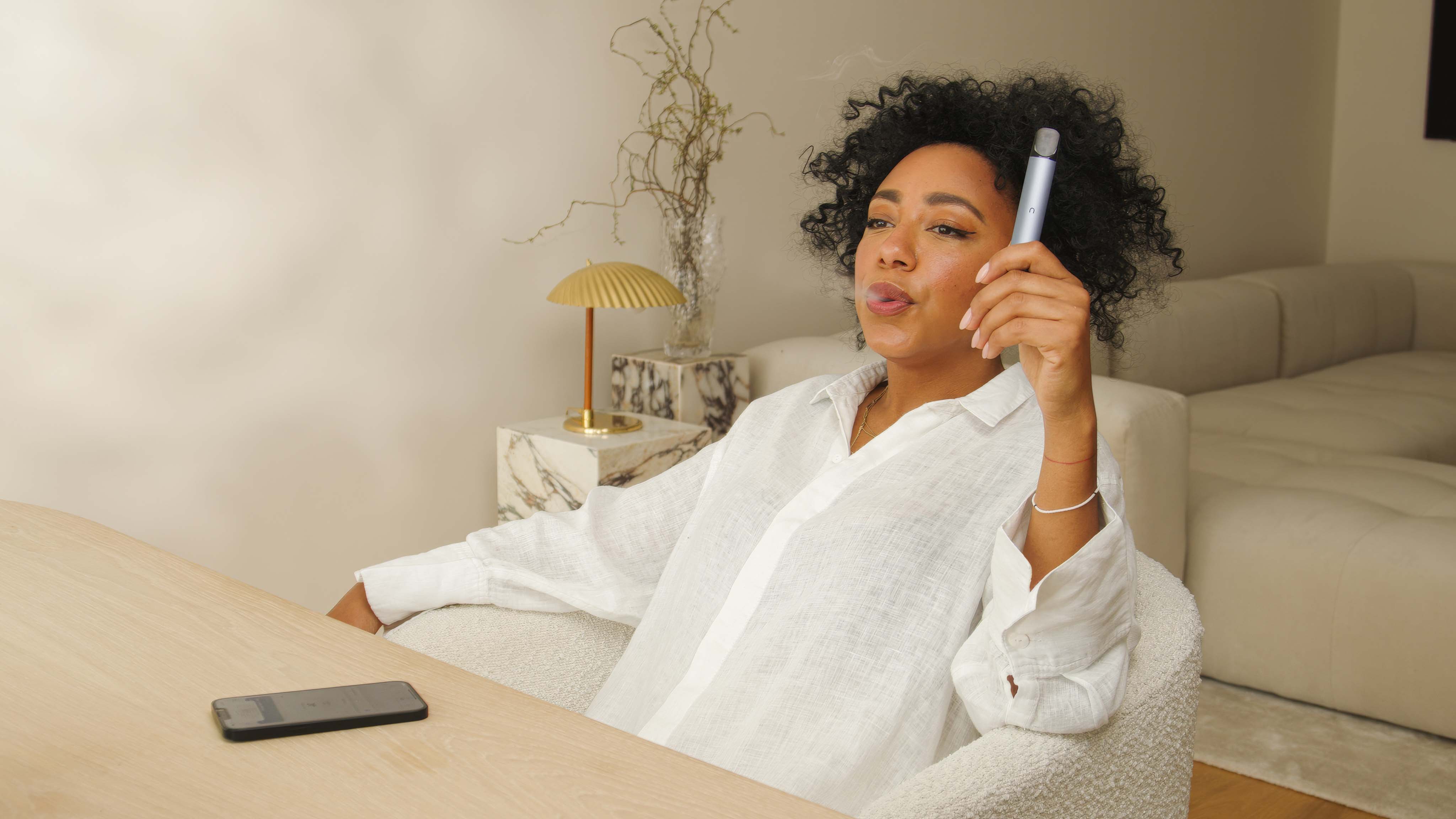 Eine Frau sitzt entspannt in einem beigen Sessel und hält einen nuumi-Vaporizer in der Hand. Auf dem Holztisch vor ihr liegt ein Smartphone, während im Hintergrund eine minimalistische Einrichtung mit einem Beistelltisch und einer Designerlampe sichtbar ist.